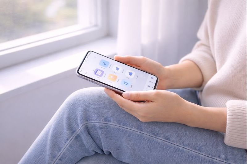 a person holding a phone loosely, sitting by a window, neutral tones